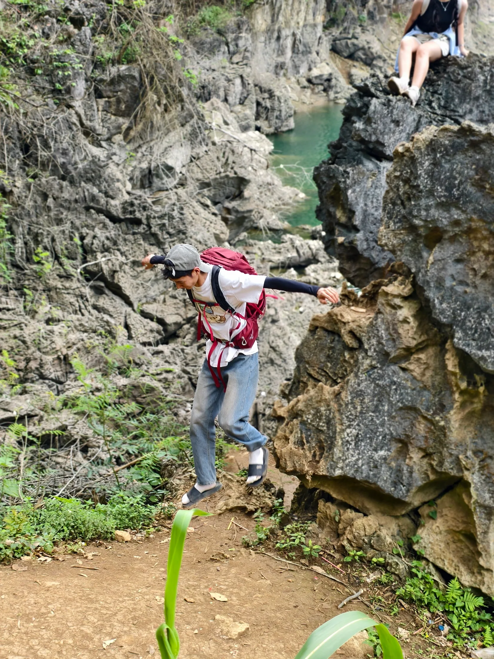 Nam du khách đeo balo đỏ nhảy qua khe đá trên vách núi, một người ngồi quan sát phía trên