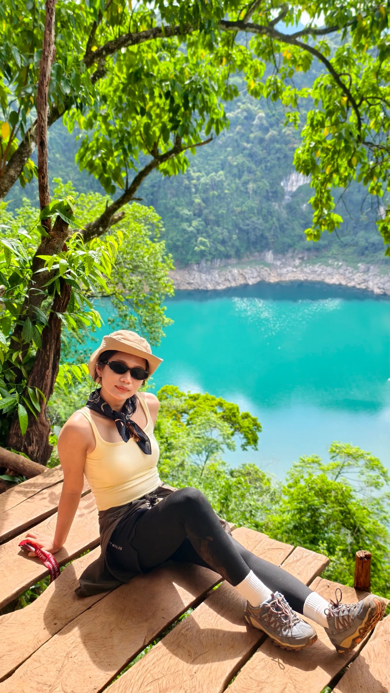 Nữ du khách ngồi nghỉ trên sàn gỗ dưới tán cây, view hồ nước xanh ngọc