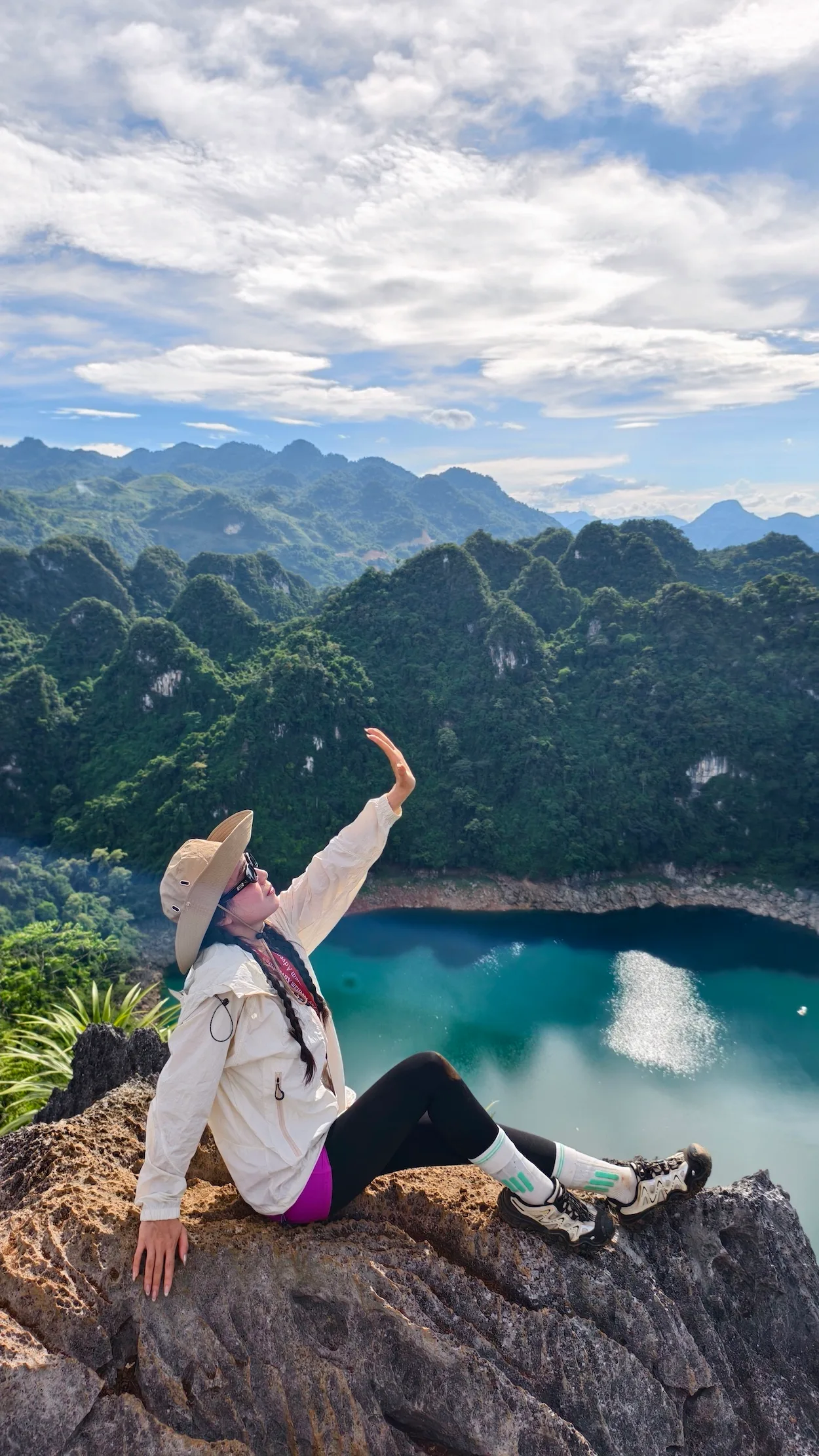 Nữ du khách ngồi trên mỏm đá giơ tay lên cao, phía sau là hồ nước và núi đá vôi