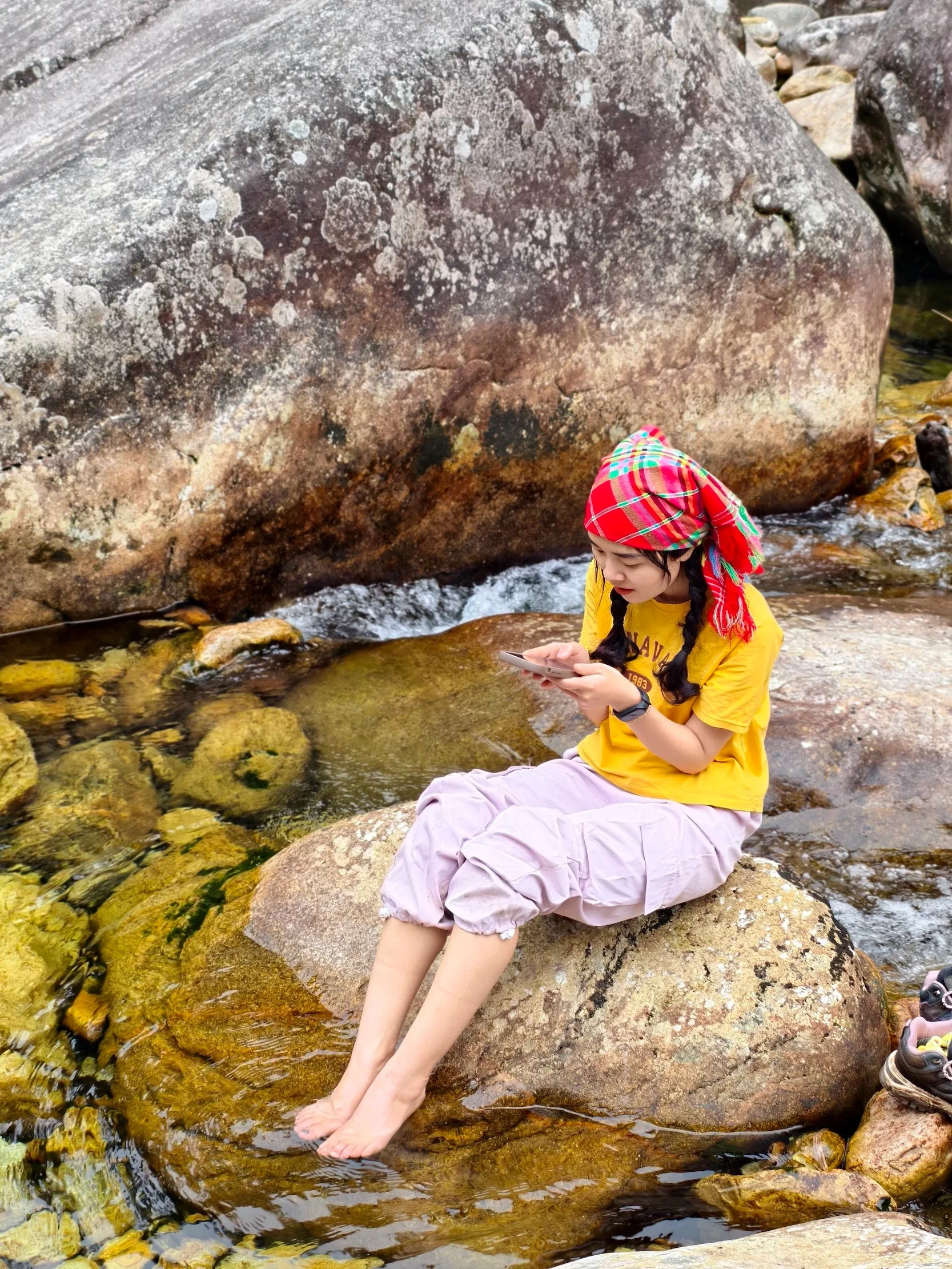 Nữ du khách áo vàng ngồi nghỉ trên đá bên dòng nước trong